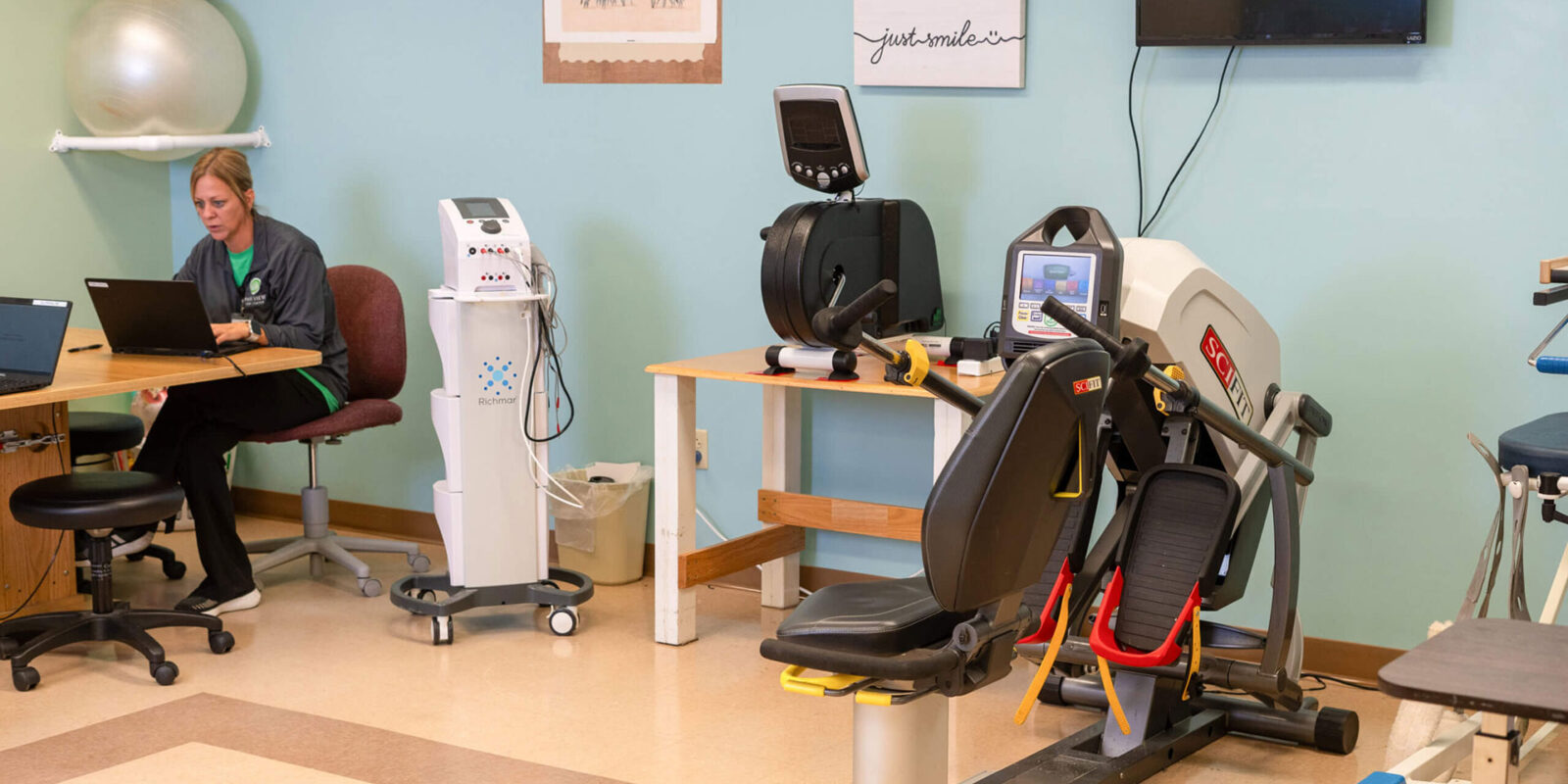 Therapy room with equipment and a person working on laptop.